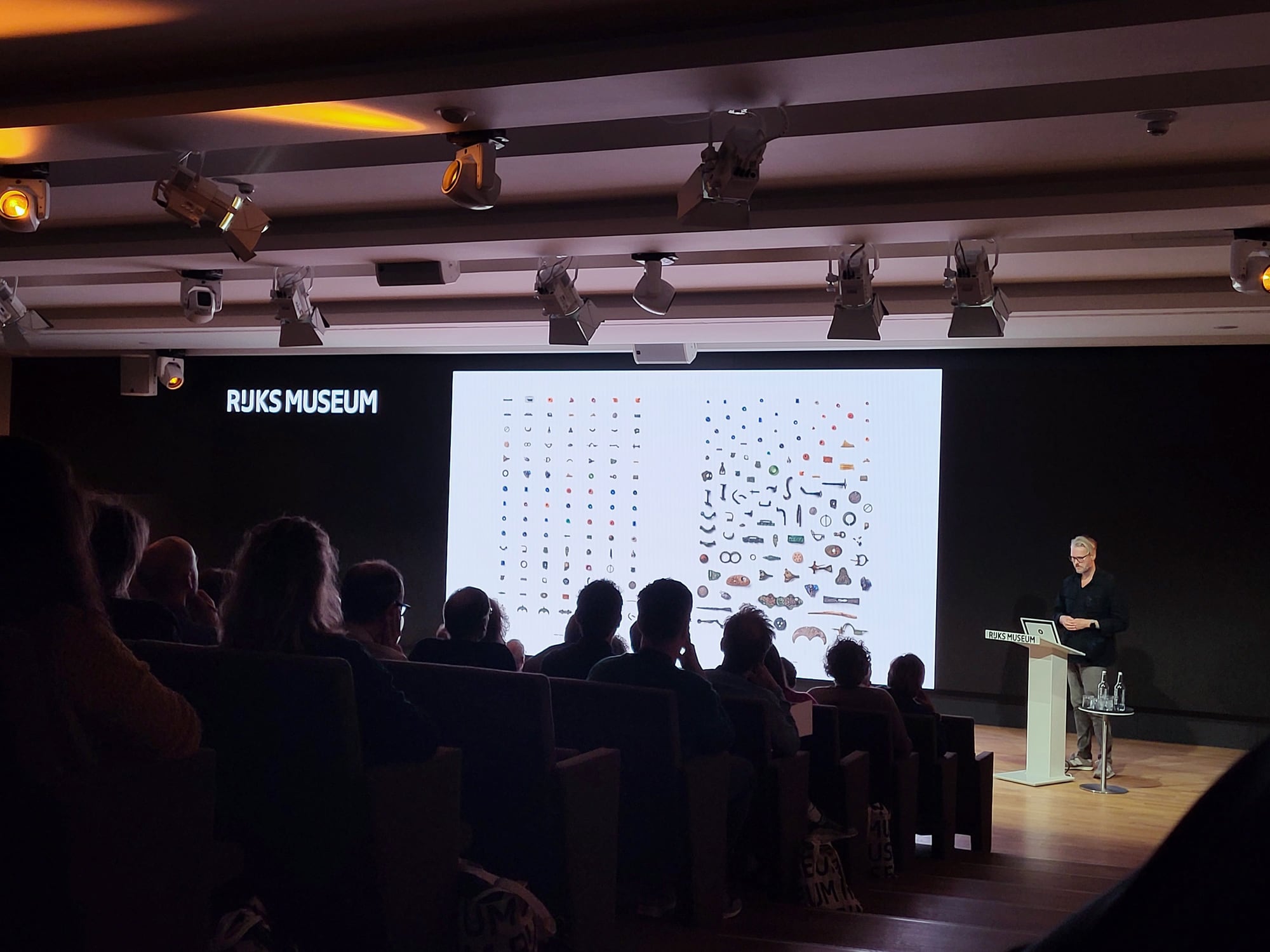 A darkened auditorium at the Rijksmuseum: the audience in silhouette, a large screen showing two grids of archaeological objects on white, and the speaker at a lectern to the right
