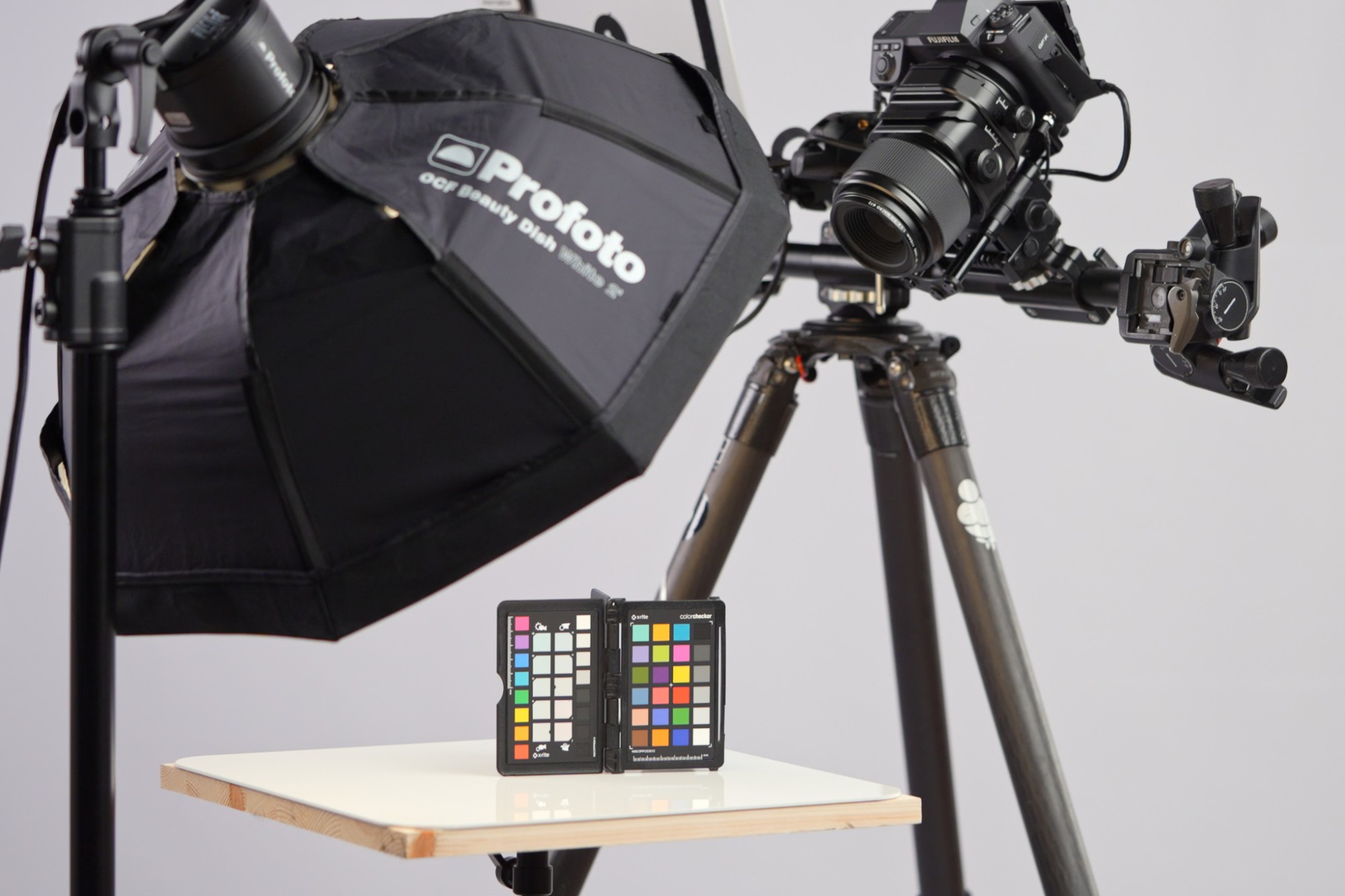 A studio setup: a beauty dish on a stand at the upper left, a camera on a geared head and arm over a tripod, and a colour card standing on a white board