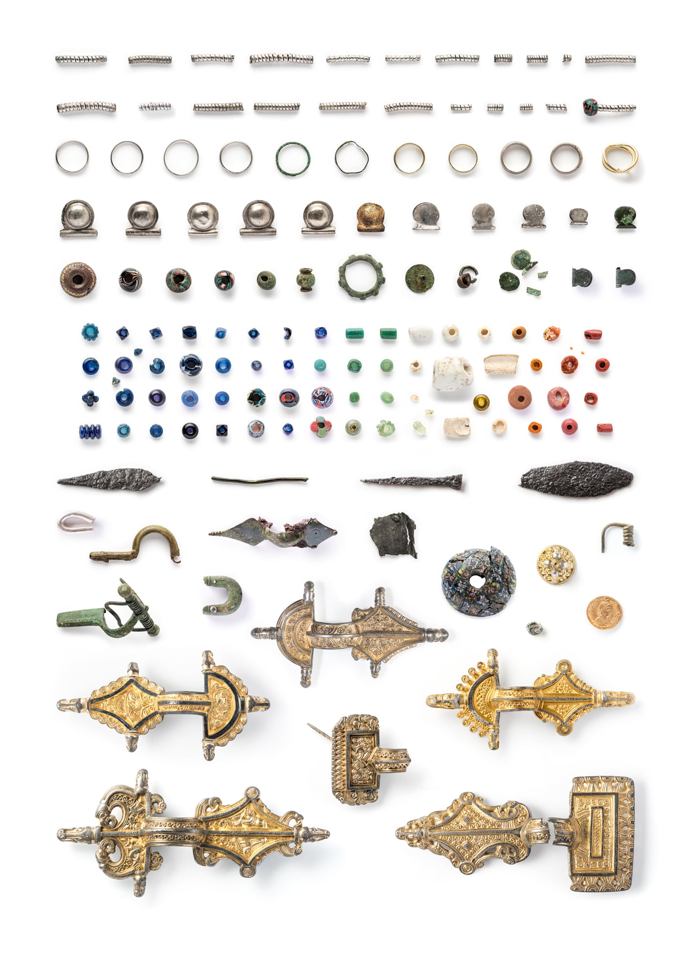 Objects from Sandby borg laid out on white in rows: spiral rings, finger rings, silver beads, glass beads in many colours, iron knives and fittings, and gilded brooches at the bottom