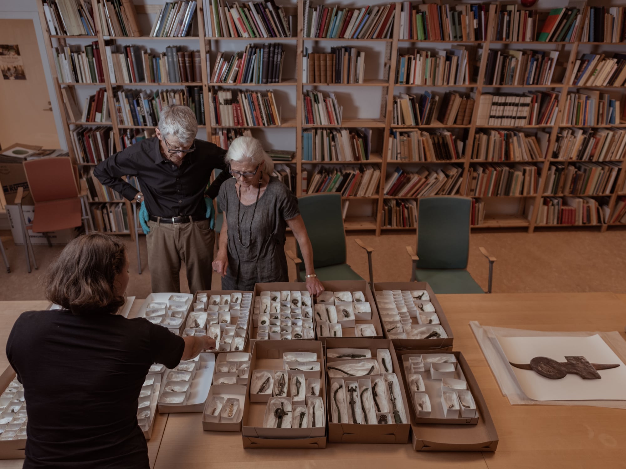 Seen from above, a table covered with boxes of the Sösdala objects in white trays; Ulf Näsman and Charlotte Fabech stand at the far side looking at them, in a room lined with bookshelves